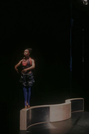 A dancer stands on a curved platform, her posture reminiscent of a classical ballet pose, surrounded by a soft glow of light.
