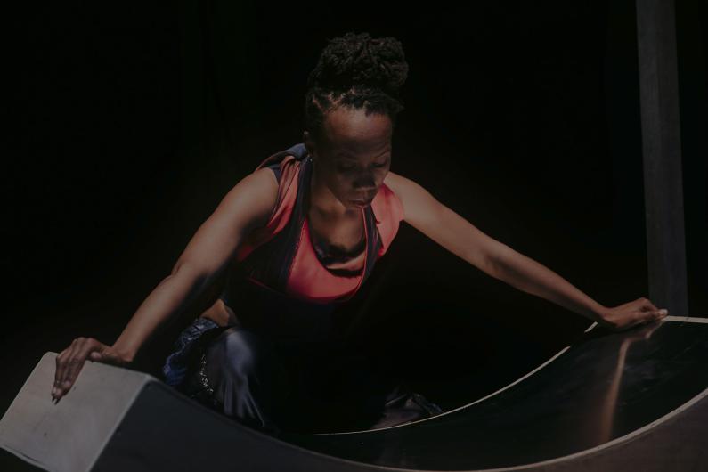 A dancer leans forward, supporting herself with her hands on a curved surface, her gaze focused, as light accentuates her facial features.