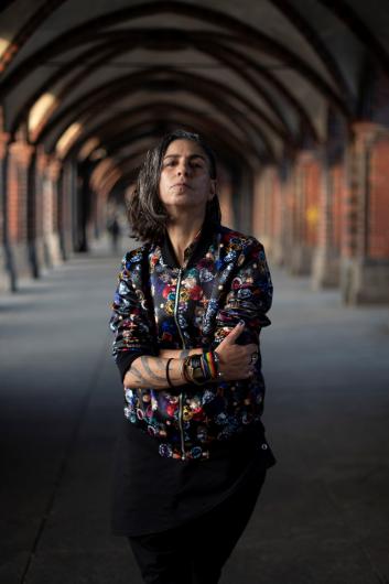 A person with medium-length dark hair stands with crossed arms in a historic archway. They wear a colorful patterned jacket and bracelets. Their gaze is confident and direct.