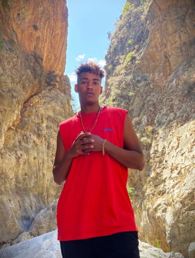 A young person with curly hair wears a red sleeveless shirt and stands between towering rock walls. The sky is blue, and the sunlight highlights the natural surroundings.