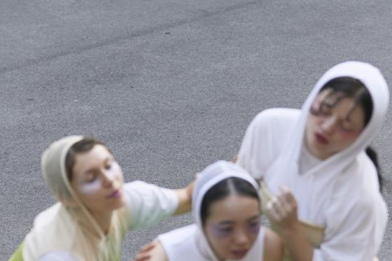 Three people with white face paint and head coverings are captured in a soft, blurred motion. Their faces are delicately painted with pastel tones around the eyes. The focus of the image highlights their dynamic movement and interaction.