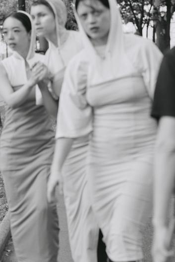 A black-and-white image shows a group of people in long, fitted dresses with hoods walking together along a pathway. Their movements appear synchronized and ceremonial.
