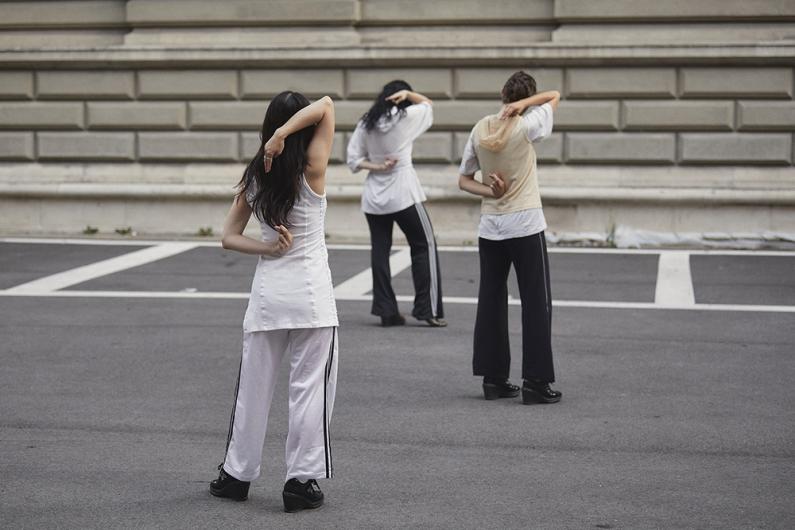 Three people stand on an empty street in front of a historic building, facing away from the camera. They wear sporty, minimalist outfits in white and beige and perform synchronized arm movements behind their backs.