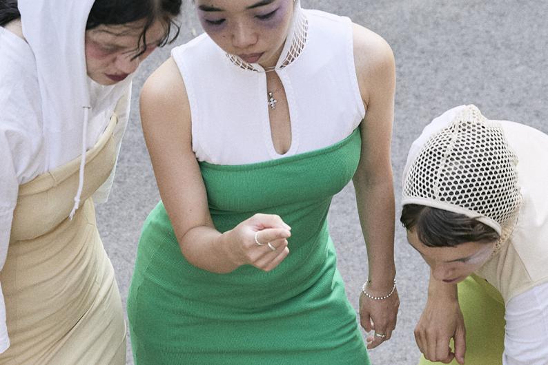 Three people in color-coordinated outfits - green, beige and white - lean forward and look at something that appears to be outside the picture in front of them.