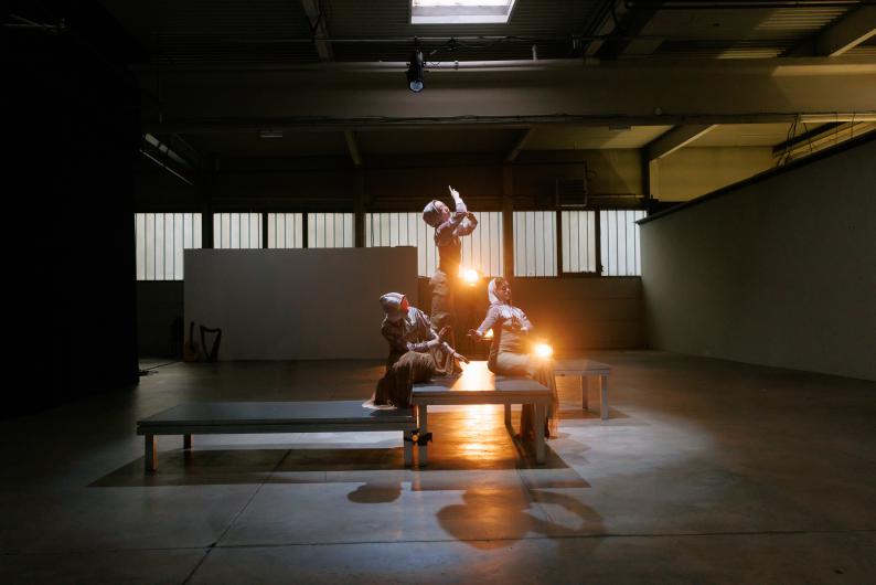 Dancers pose on tiered platforms in an industrial space, lit by dramatic stage lighting.