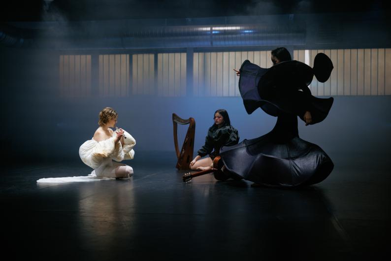 Three performers in elaborate outfits, flanked by a harp and guitar, in a mysteriously lit scene.