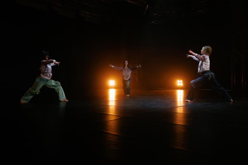 Three dancers in a dramatic pose on a dark stage, illuminated by warm lights in the background.
