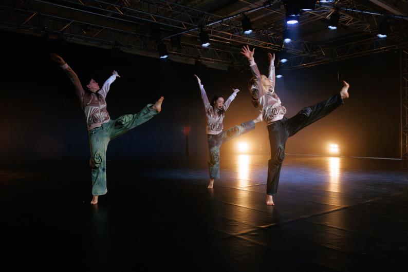 Three dancers with raised arms and outstretched legs in a synchronized movement on stage.