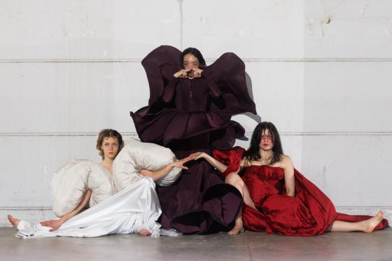 Three dancers in elaborate costumes – white, purple, and red – pose artfully in front of a white wall.