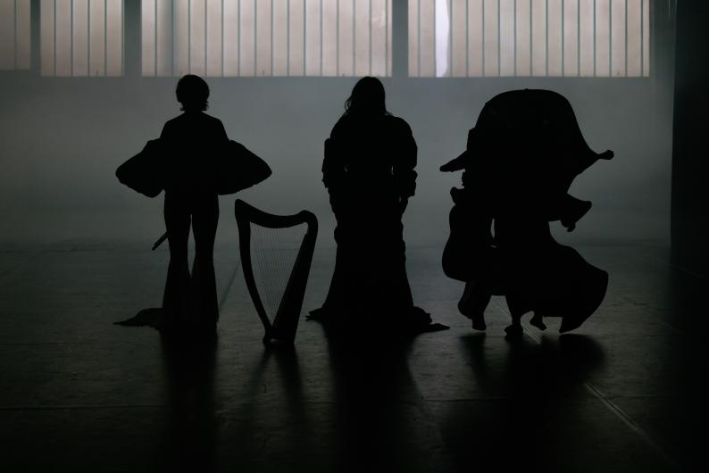 Silhouettes of three performers with instruments backlit in a foggy, dramatically lit space.