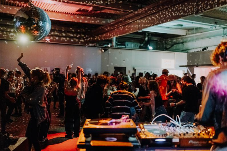 A vibrant party scene in a club or event space. In the foreground, a DJ booth with turntables and mixers is visible, while a dancing crowd celebrates in the background under a disco ball. The reflections from the disco ball cast sparkling dots on the walls and ceiling. The atmosphere appears lively and energetic.