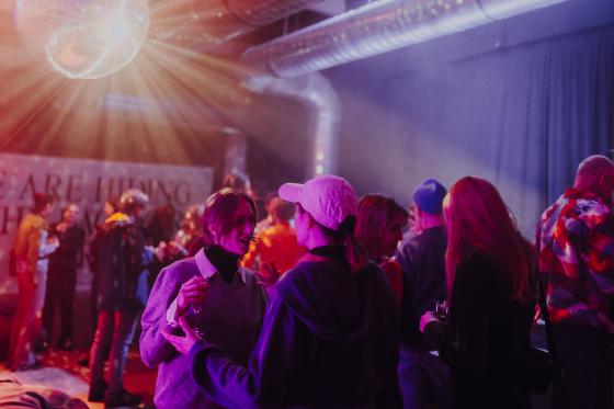 Eine Gruppe junger Menschen unterhält sich bei einer Party oder Veranstaltung in einem Club mit lila und rotem Licht. Eine Discokugel strahlt Lichtstrahlen aus der Decke, während die Gäste Getränke in der Hand halten und in entspannter Atmosphäre plaudern. Im Hintergrund ist ein Transparent mit dem Text „WE ARE HIDING ...“ teilweise sichtbar.