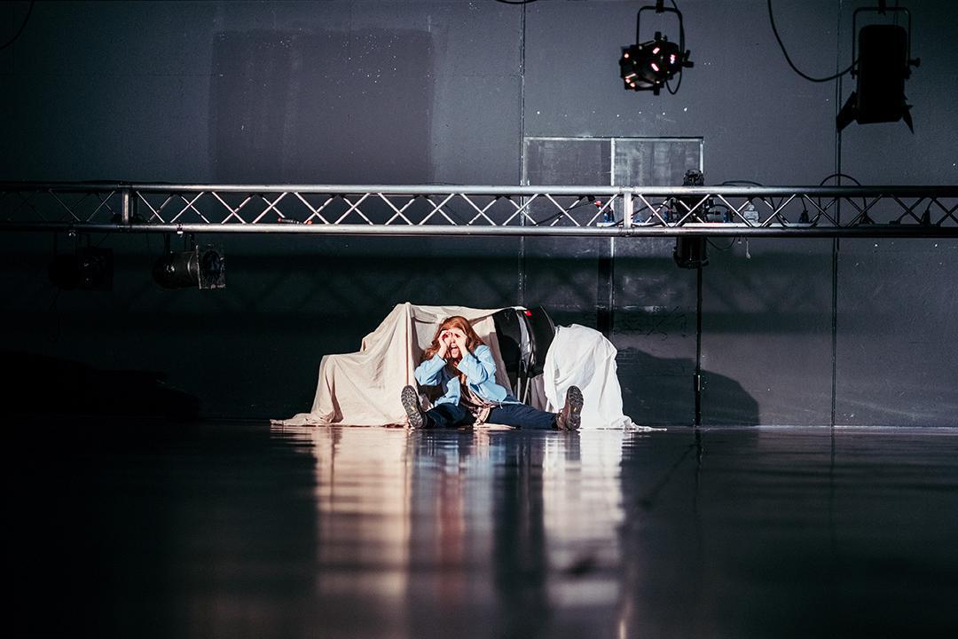 A person is seated on the floor of a dark performance space, surrounded by stage equipment and white fabric draped over an object. The person is wearing a light blue top and dark pants, holding their face with both hands, as if in thought or distress. Overhead, lighting rigs are visible.