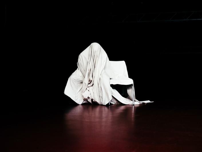 A dark stage with a person hidden under a large white cloth. The fabric drapes over a chair and the floor, creating a mysterious and ghostly atmosphere.
