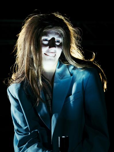 A close-up of a person with dramatic lighting illuminating their face, casting shadows that create an eerie effect. They are wearing a blue blazer, and their long hair is disheveled. Their wide eyes and slightly exaggerated smile give a haunting and unsettling expression.
