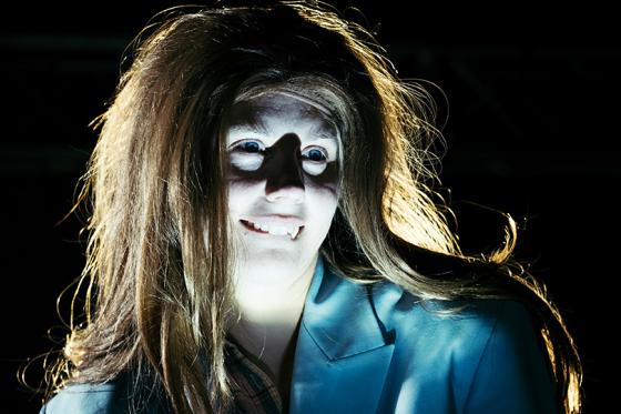 Eine Nahaufnahme einer Person mit dramatischer Beleuchtung, die ihr Gesicht erhellt und Schatten wirft, die einen unheimlichen Effekt erzeugen. Sie trägt einen blauen Blazer, und ihr langes Haar ist zerzaust. Ihre weit geöffneten Augen und das leicht übertriebene Lächeln verleihen ihr einen gespenstischen und beunruhigenden Ausdruck.