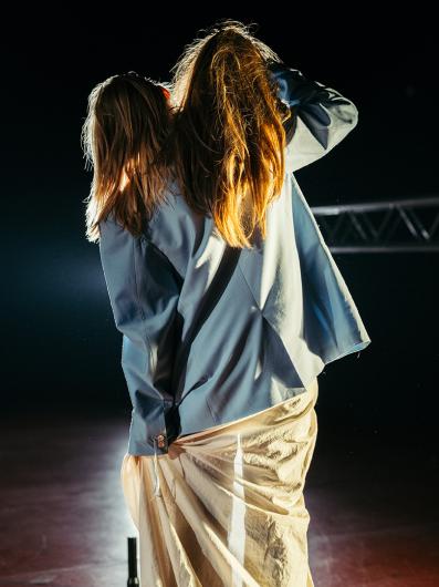A person with long hair, seen from behind, wearing a blue jacket and a long beige fabric skirt. She holds a wig in her hand and therefor it almost looks like she hast wo heads The lighting emphasizes movement and the textures of the clothing.