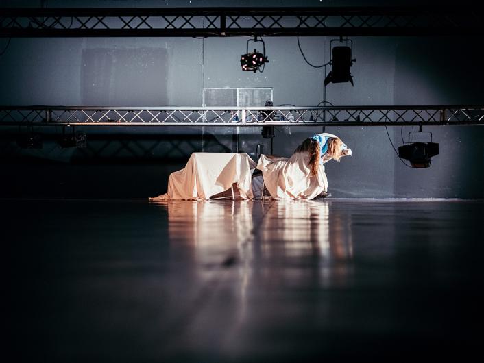 A person is seated on the floor of a dark performance space, surrounded by stage equipment and white fabric draped over an object. The person is wearing a light blue top and dark pants, holding their face with both hands, as if in thought or distress. Overhead, lighting rigs are visible.
