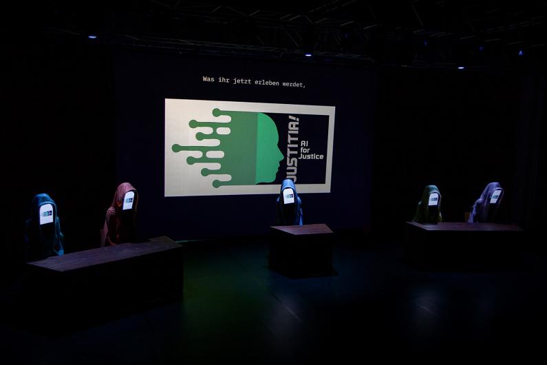 A dark stage with four figures wearing hoods and having screens as faces. In the background, a projection with the title “JUSTITIA! AI for Justice” and an abstract profile image in green. The text at the top reads: “Was ihr jetzt erleben werdet” ("What you are about to experience").