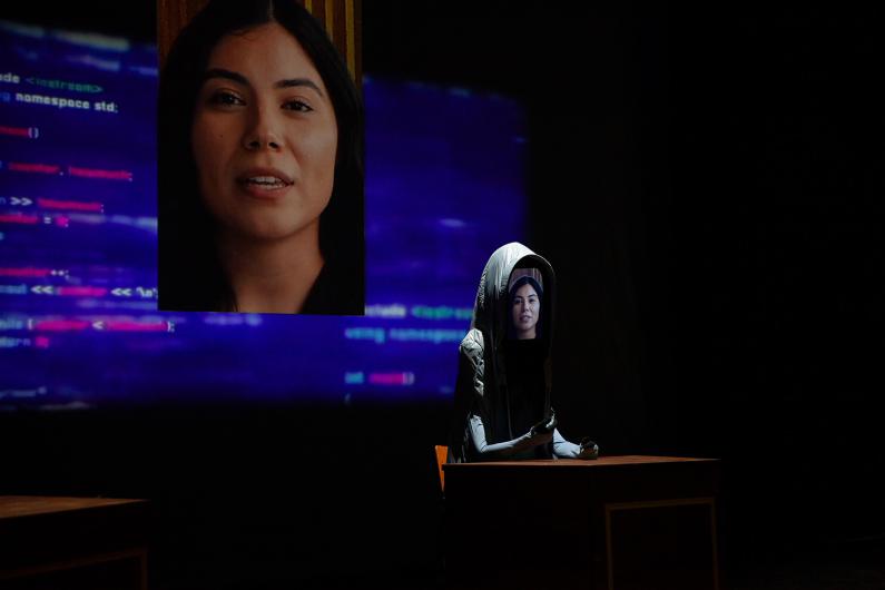 Eine Figur mit Kapuze sitzt an einem Tisch. Auf dem Bildschirm ihres „Gesichts“ wird eine Frau gezeigt. Im Hintergrund ist ein großer Bildschirm mit dem gleichen Gesicht und digitalen Codezeilen in lila-blau zu sehen.