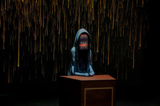 A hooded figure sits at a wooden desk. Its “face” displays colorful vertical data lines with a red horizontal bar. In the background, a projection of golden vertical lines on a black background.