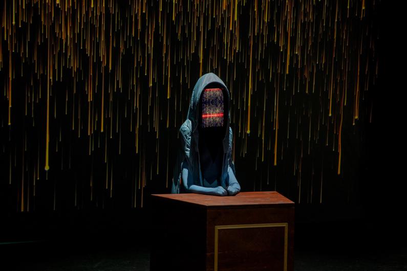 A hooded figure sits at a wooden desk. Its “face” displays colorful vertical data lines with a red horizontal bar. In the background, a projection of golden vertical lines on a black background.