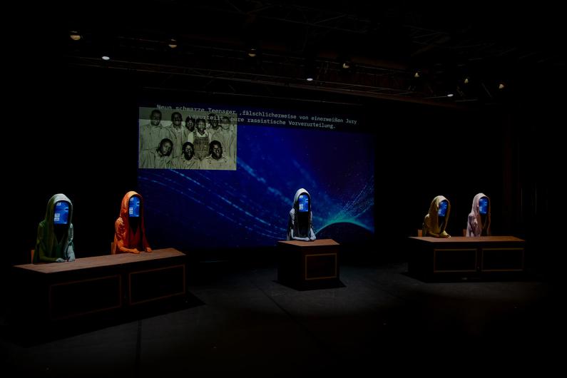 Five hooded figures sit at desks in a dimly lit room. Their faces are replaced by screens emitting a blue glow. A historical black-and-white image is projected in the background, accompanied by German text about racial prejudice and wrongful convictions of black teenagers.