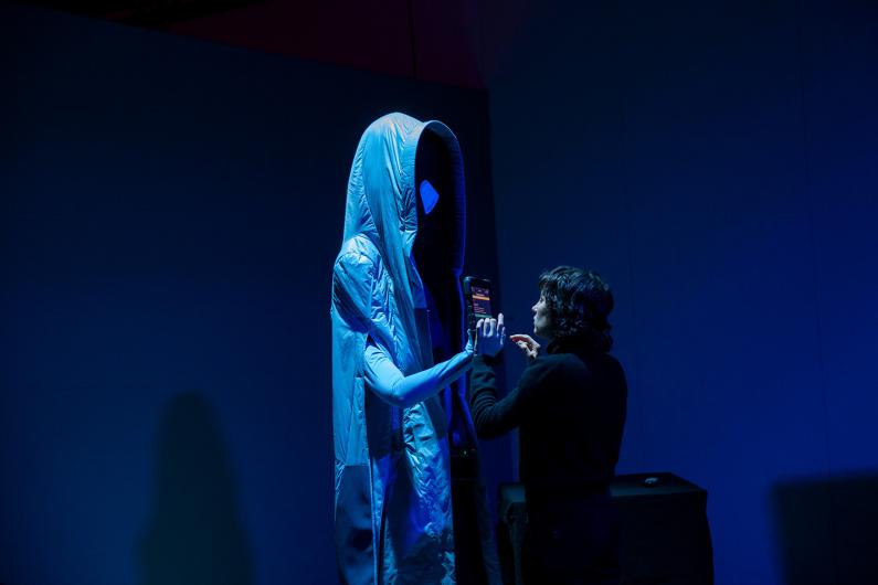 Eine Person steht vor einer großen, humanoiden Figur mit einer blauen, futuristischen Kapuze und einem ovalen Gesicht ohne erkennbare Merkmale. Sie hält ein Gerät und interagiert mit der Figur in einem schwach beleuchteten Raum in blauem Licht.