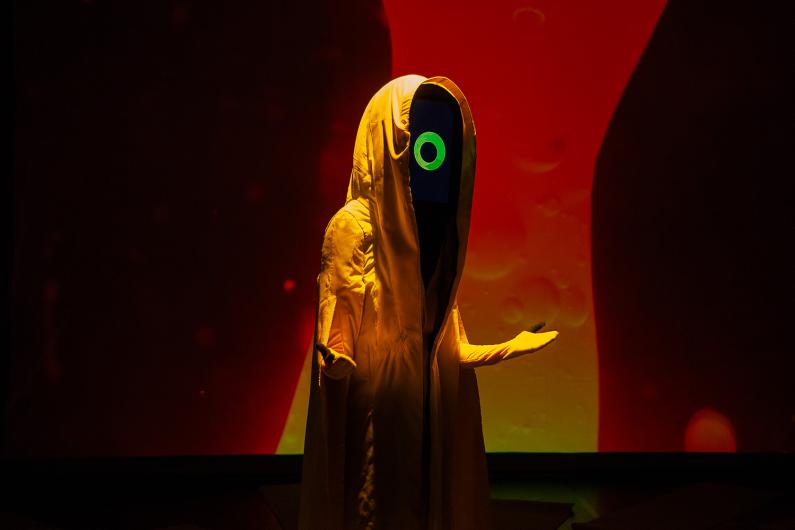 A hooded figure in a yellow robe stands with its arms outstretched. The figure’s face is represented by a black screen displaying a green circular symbol. The background is lit in intense red and orange hues.