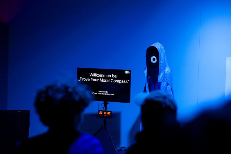 Eine humanoide Figur in einem futuristischen blauen Gewand steht neben einem Bildschirm mit der Aufschrift „Willkommen bei ‚Prove Your Moral Compass‘“. Zuschauer betrachten die Szene in einem Raum mit blauem Licht.