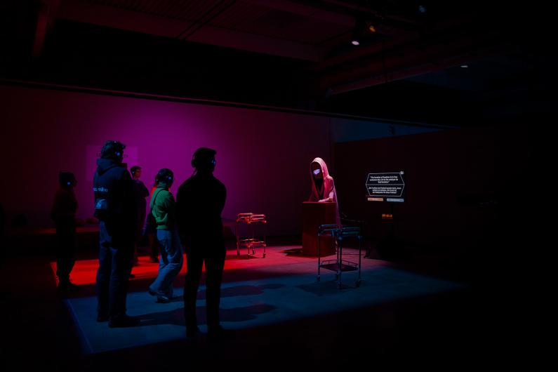 A single hooded figure with a glowing screen for a face stands behind a podium, illuminated by purple and red lights. Visitors wearing headphones observe the scene, while text is projected onto a screen to the right.