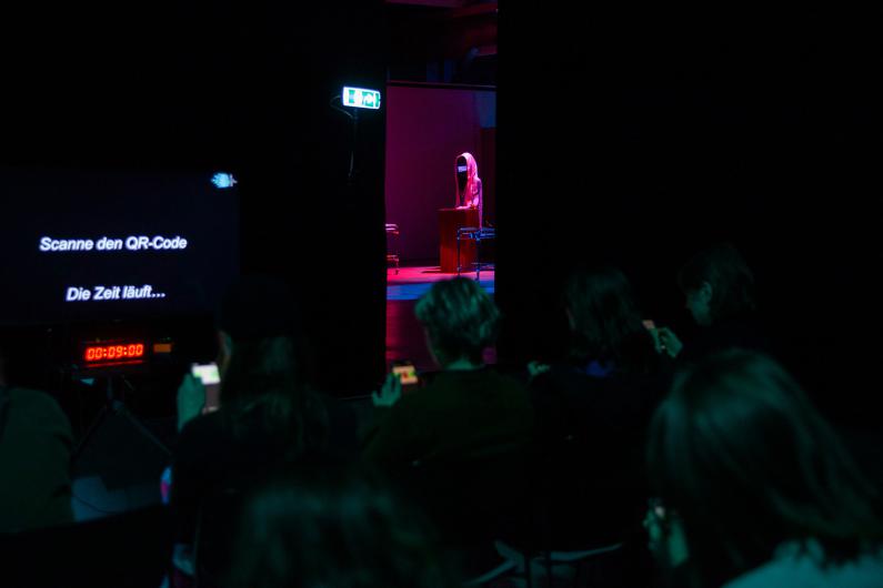 A view through a dark room reveals a hooded figure at a podium bathed in purple light. In the foreground, audience members sit with smartphones, while a screen to the left displays a QR code and a countdown timer.
