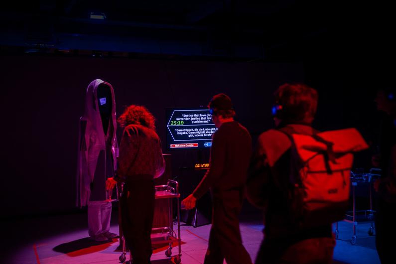 A group of people gathers around a hooded figure with a black screen for a face. A nearby monitor displays text attributed to Mahatma Gandhi, with the scene illuminated in dim red light.