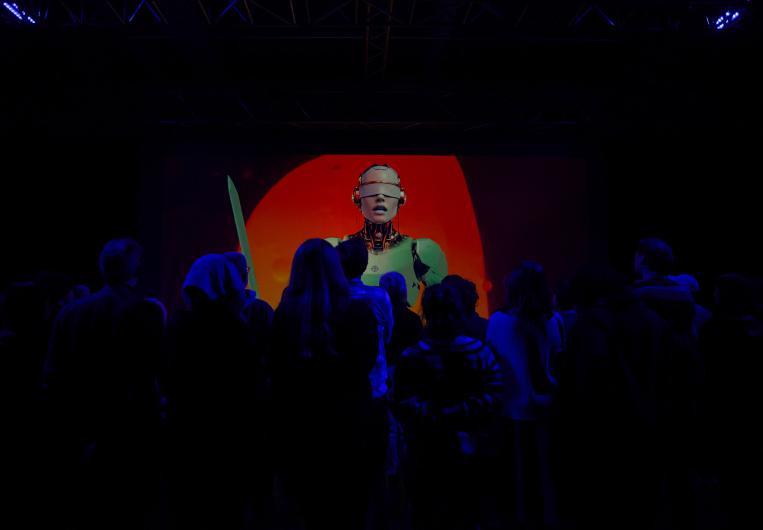 A group of spectators stands in front of a large screen displaying an android figure with a futuristic appearance and a sword against a red background.