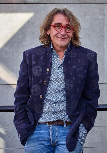 Ein mittelalter Mann mit schulterlangem welligem Haar und roten Brillen, trägt einen gemusterten dunkelblauen Blazer, ein hellblau gemustertes Hemd und eine blaue Jeans. Er steht entspannt vor einer Betonwand und einem Geländer.