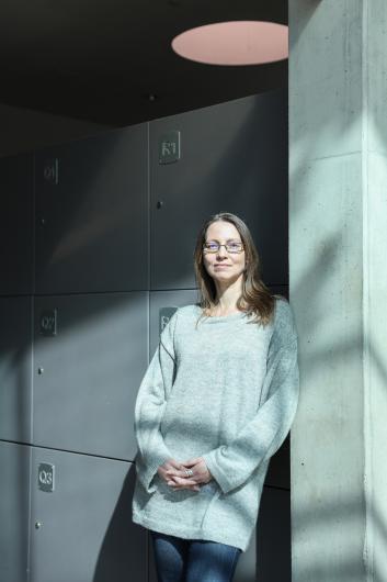 Eine Frau mit Brille und einem hellgrauen Pullover lehnt an einer Betonsäule und hat die Hände vor sich gefaltet. Sie befindet sich in einem modernen Innenraum mit grauen Schließfächern im Hintergrund, und Sonnenlicht wirft sanfte Schatten an die Wand.