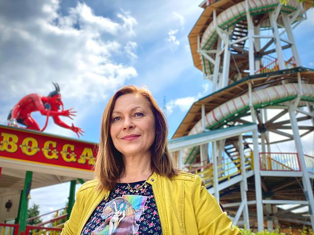 A smiling woman in a yellow jacket stands in front of an amusement park attraction on a sunny day. Behind her, there is a tall spiral slide and a red devil statue on a sign that reads "BOCCA." The sky is partly cloudy.