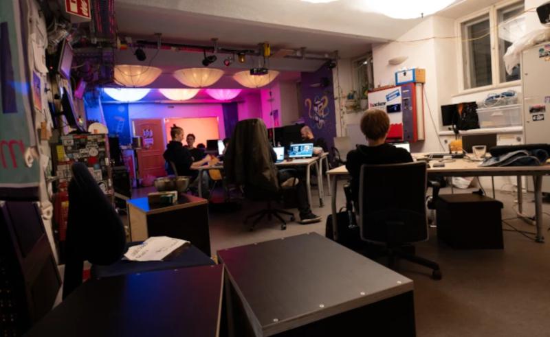 Ein gemeinschaftlicher Arbeitsbereich mit mehreren Personen, die an Schreibtischen sitzen und an Laptops arbeiten. Der Raum ist schwach beleuchtet, mit farbiger Ambientebeleuchtung in Rosa- und Blautönen, und die Wände sind mit Postern und kreativer Dekoration geschmückt. Auf der rechten Seite sind ein Getränkeautomat und Regale mit verschiedenen Gegenständen zu sehen.