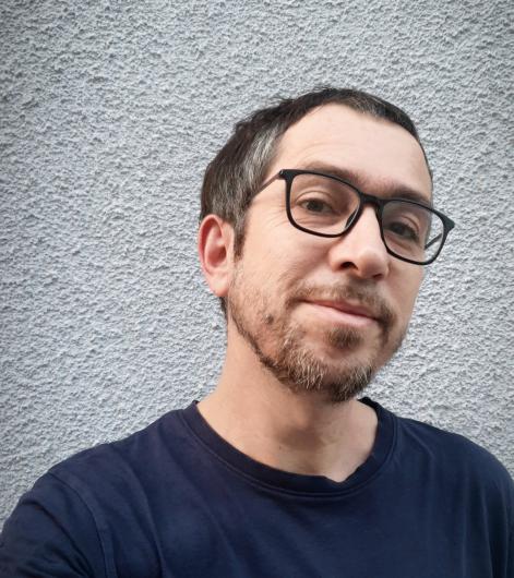 A close-up of a man with short dark hair and gray highlights wearing rectangular glasses and a navy blue t-shirt, standing in front of a textured light gray wall. He is smiling slightly with a relaxed expression.