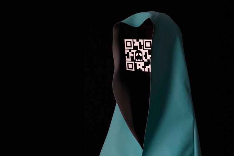 Ein digital stilisiertes Objekt, das einer in blaugrünfarbenem Stoff gehüllten Figur ähnelt, mit einem schwarzen, bildschirmartigen Gesicht, das einen QR-Code zeigt. Der QR-Code enthält ein kleines, geisterförmiges Muster in seinem Design. Das Bild ist vor einem dunklen Hintergrund aufgenommen, was das futuristische und geheimnisvolle Aussehen des Objekts betont.