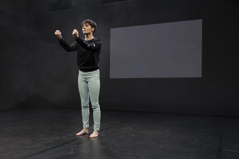 Eine Performerin steht barfuß auf einer dunklen Bühne, trägt einen schwarzen Pullover und hellgrüne Hosen. Ihre Arme sind erhoben, die Hände gestikulieren. Ein leeres weißes Rechteck ist an der Wand im Hintergrund zu sehen.