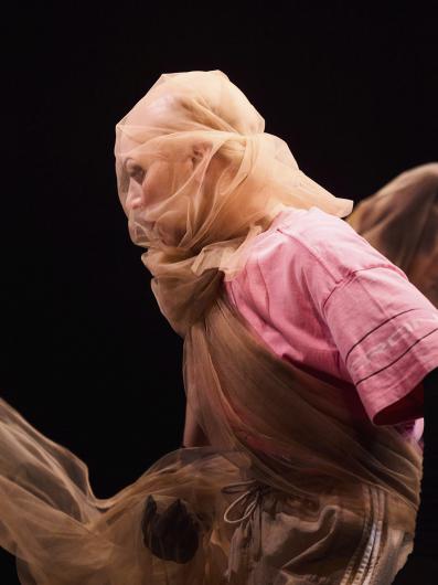 A person with their head and upper body wrapped in sheer, beige fabric that obscures their face and adds a flowing, ethereal effect. They are wearing a loose pink shirt and beige pants with a drawstring, creating a soft, layered look. The background is dark, bringing focus to the fabric's texture and the subtle contours of the person's face beneath the translucent material.