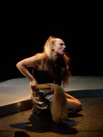 Ein*e Performer*in mit langem, dunkelbraunem und blondem Haar kniet auf der Bühne bei gedämpftem Licht und hält einen hellbraunen Stoff in einer ausdrucksstarken Pose mit schmerzverzerrtem Gesichtsausdruck.