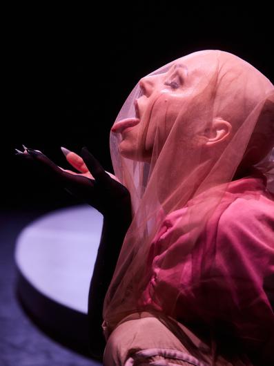 Eine Person mit Glatze und transparentem Schleier, die ihre Zunge herausstreckt. Die Person trägt schwarze Handschuhe, und der Gesichtsausdruck wirkt ekstatisch oder leidenschaftlich. Der Schleier verstärkt die theatrale Wirkung.