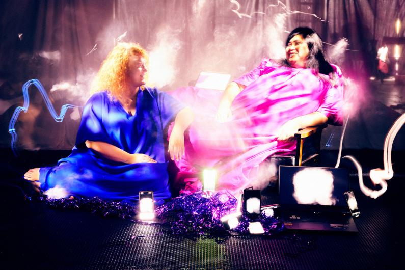 Two individuals sit in a surreal setting with dramatic lighting and fog. The person on the left wears a bright blue dress, while the one on the right wears a pink dress. Around them are scattered tech devices like smartphones and a laptop, some of which are glowing. The scene appears futuristic and artistic.