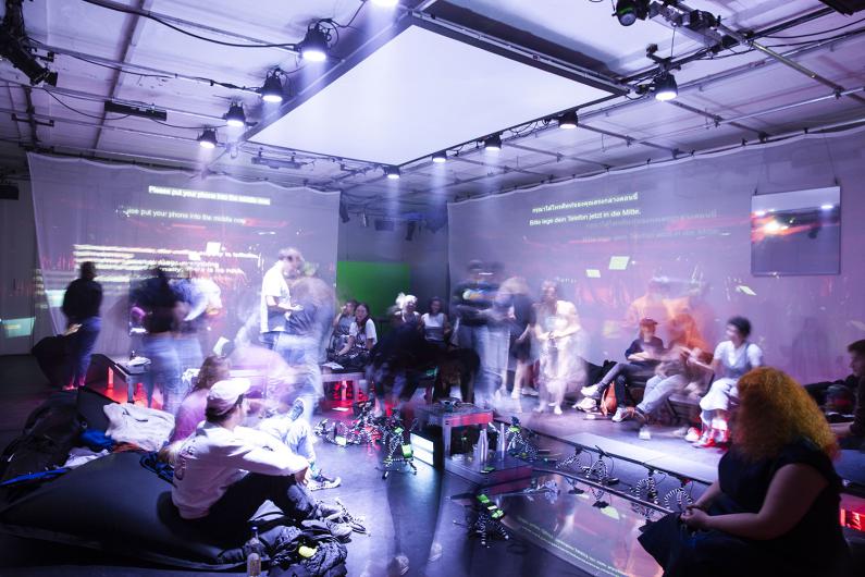 A dark room with several people sitting on benches and cushions scattered across the space. Bright, radial light projections are visible on the walls and ceiling. The floor reflects the colorful light of the installations. Some people are looking at their smartphones, while others are observing the light projections.