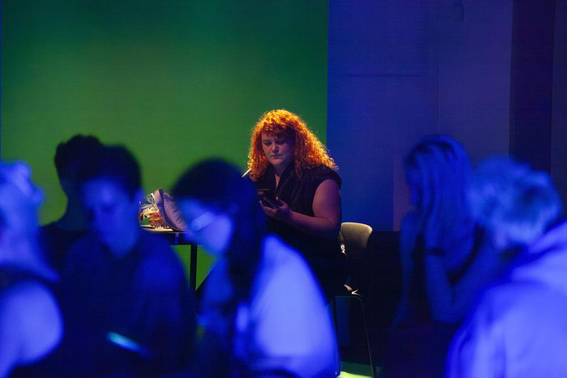 A person with bright red hair sits focused at a table in front of a green background, looking at their smartphone. In the foreground, blurred figures in blue light add depth to the scene.