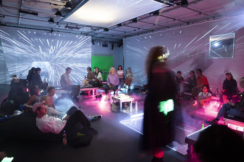 A dark room with several people sitting on benches and cushions scattered across the space. Bright, radial light projections are visible on the walls and ceiling. The floor reflects the colorful light of the installations. Some people are looking at their smartphones, while others are observing the light projections.