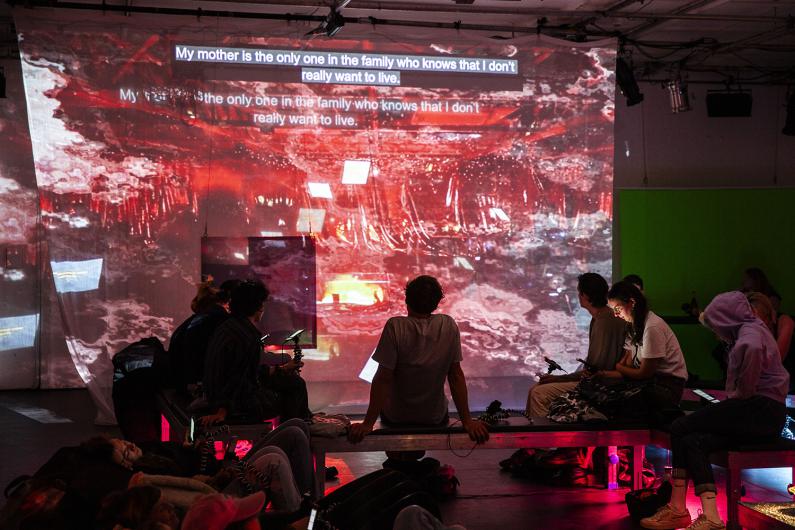 A group of people sits in front of a wall with a large-scale projection in red and brown hues. A caption at the top reads: “My mother is the only one in the family who knows that I don’t really want to live.” Some individuals are seated on benches, while others relax on the floor.