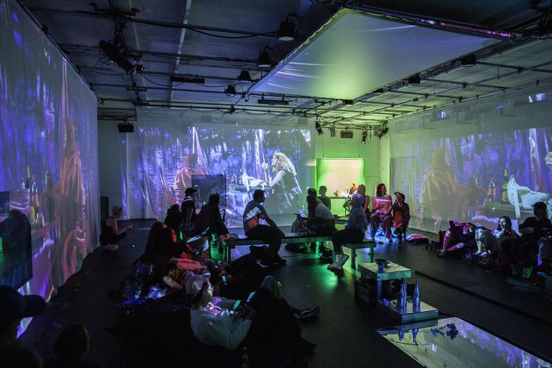 A dark room with projections in blue and green tones, displaying scenes of people at tables. The audience is seated on benches and cushions, observing the projections visible on multiple walls. Some appear to be relaxing while sitting or lying down.